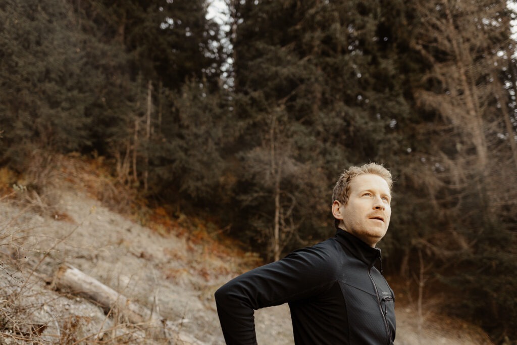 A man in a black jacket standing in the woods with mountains in the background.