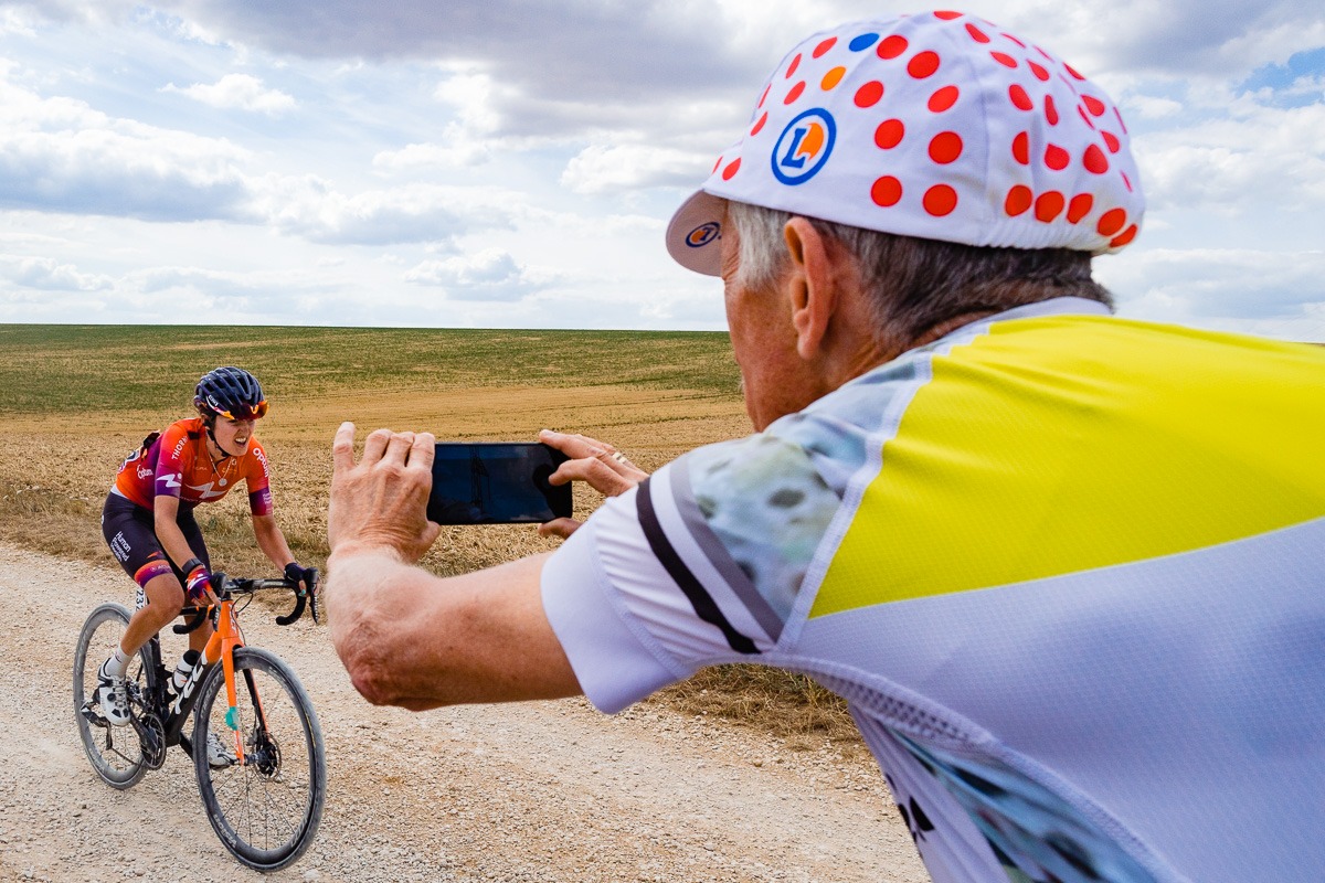 Ein Radfahrer fährt auf einer Schotterstraße, während eine andere Person mit einer gepunkteten Mütze in einer ländlichen Außenumgebung ein Foto mit einem Smartphone macht.