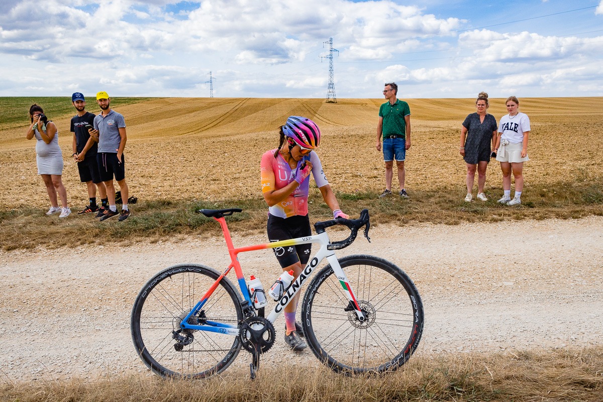 Ein Radfahrer auf einer Schotterstraße hält an, um Wasser zu trinken, während eine kleine Gruppe von Zuschauern an der Seite steht, beobachtet und Fotos macht.