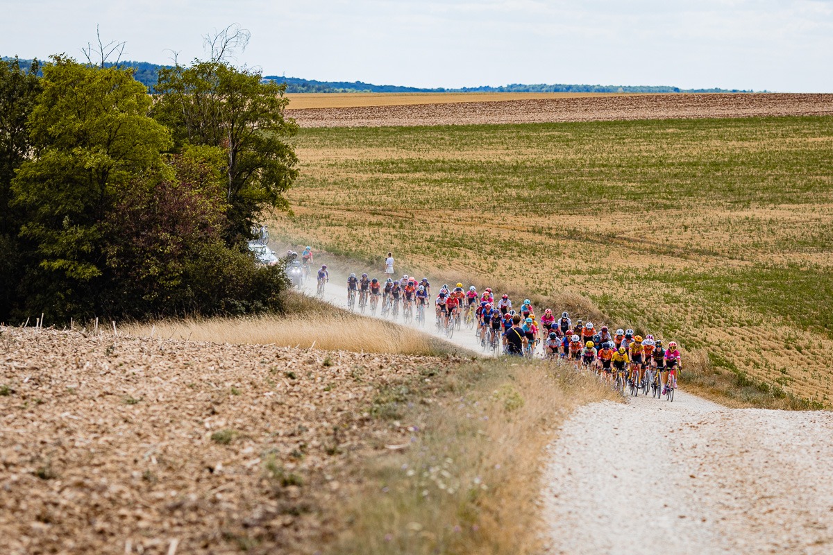 Eine Gruppe von Radfahrern fährt auf einer staubigen Landstraße, die von Feldern und Bäumen umgeben ist, unter einem teilweise bewölkten Himmel.