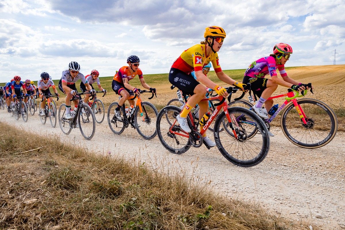 Eine Gruppe von Radfahrern in bunten Outfits fährt unter einem teilweise bewölkten Himmel auf einem Kiesweg, der von Feldern umgeben ist.