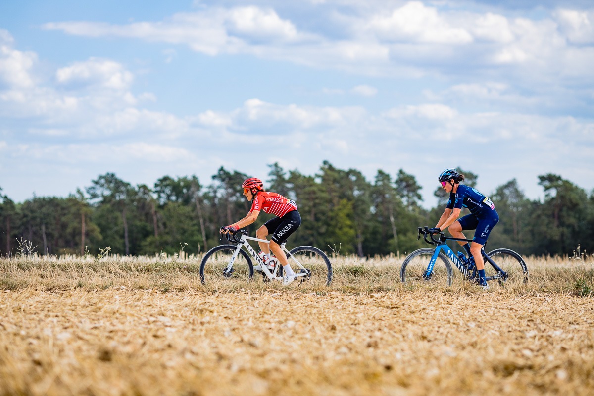Zwei Radfahrer fahren auf einem Pfad durch ein grasbewachsenes Feld unter einem teilweise bewölkten Himmel mit Bäumen im Hintergrund. Der eine trägt ein rotes und der andere ein blaues Trikot.