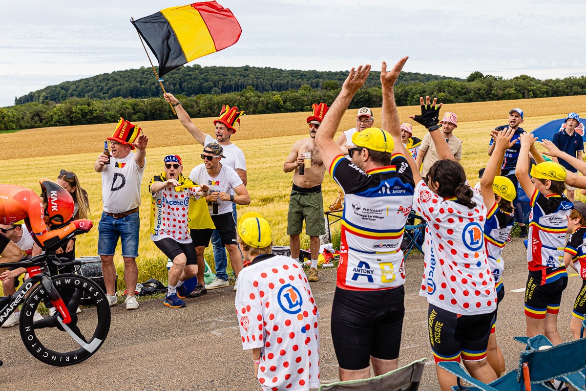 Eine Gruppe von Radsportfans, von denen viele bunte Kleidung und gepunktete Trikots tragen, feuern bei einem Rennen begeistert einen Radfahrer an, wobei einer von ihnen in einer ländlichen Landschaft eine belgische Flagge schwenkt.
