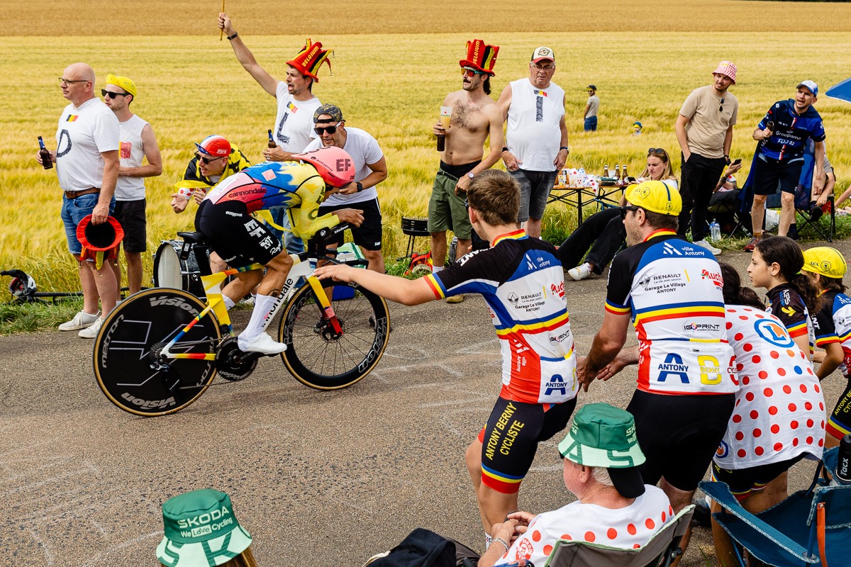 Ein Radfahrer nimmt an einem Zeitfahren teil, während Zuschauer, einige mit bunten Hüten und Anzügen, am Rande einer Landstraße vor dem Hintergrund eines Weizenfeldes applaudieren und Fotos machen.