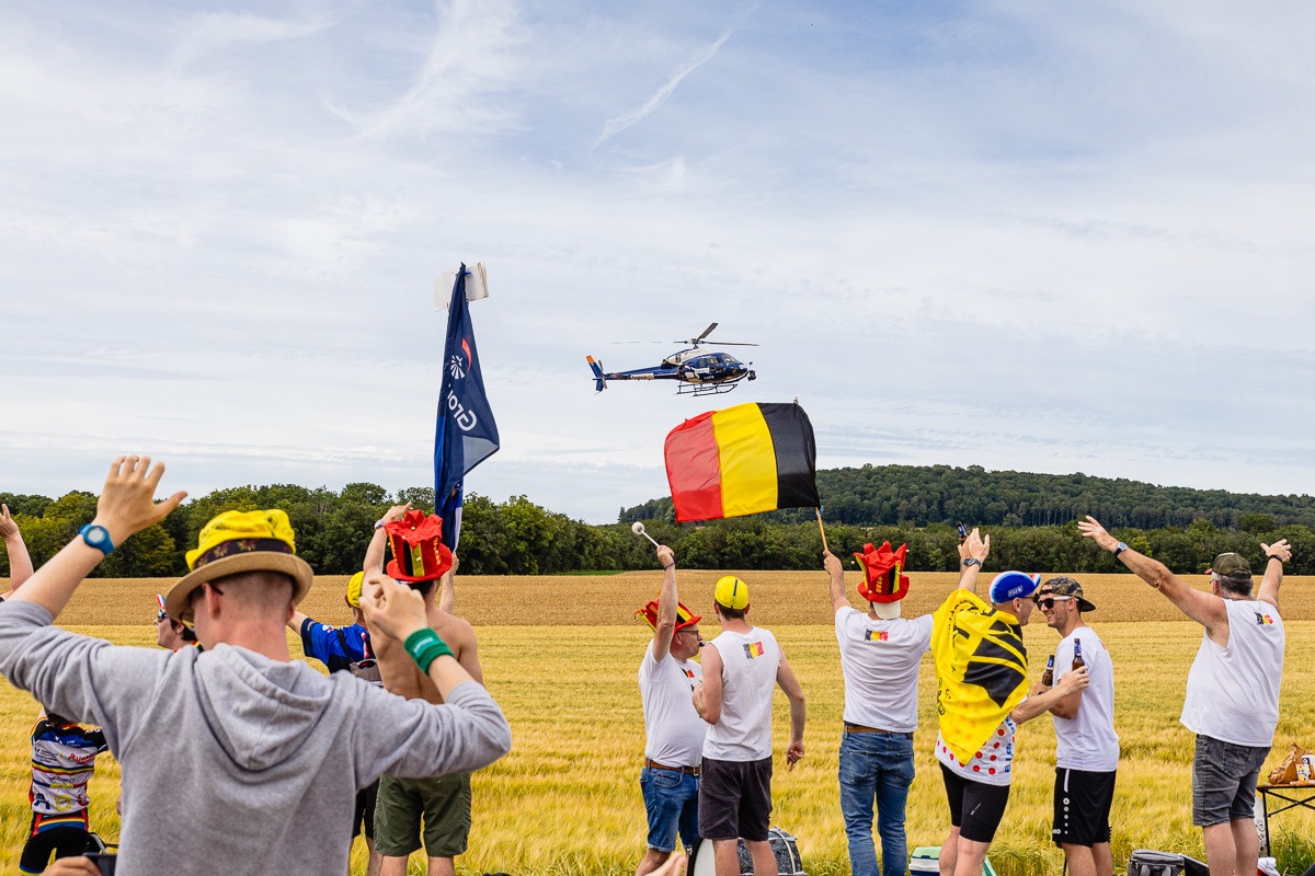 Eine Gruppe von Personen winkt einem Hubschrauber zu, der über die Bühne fliegt. Einige Personen schwenken eine belgische Flagge und tragen thematisch passende Accessoires. Sie befinden sich auf einem Feld mit einem Waldgebiet im Hintergrund.