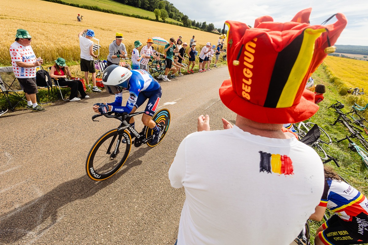 Ein Radfahrer nimmt an einem Rennen auf einer Landstraße teil und wird von Fans bejubelt, von denen einer einen großen Hut in den Farben von "Belgien" und der belgischen Flagge trägt.