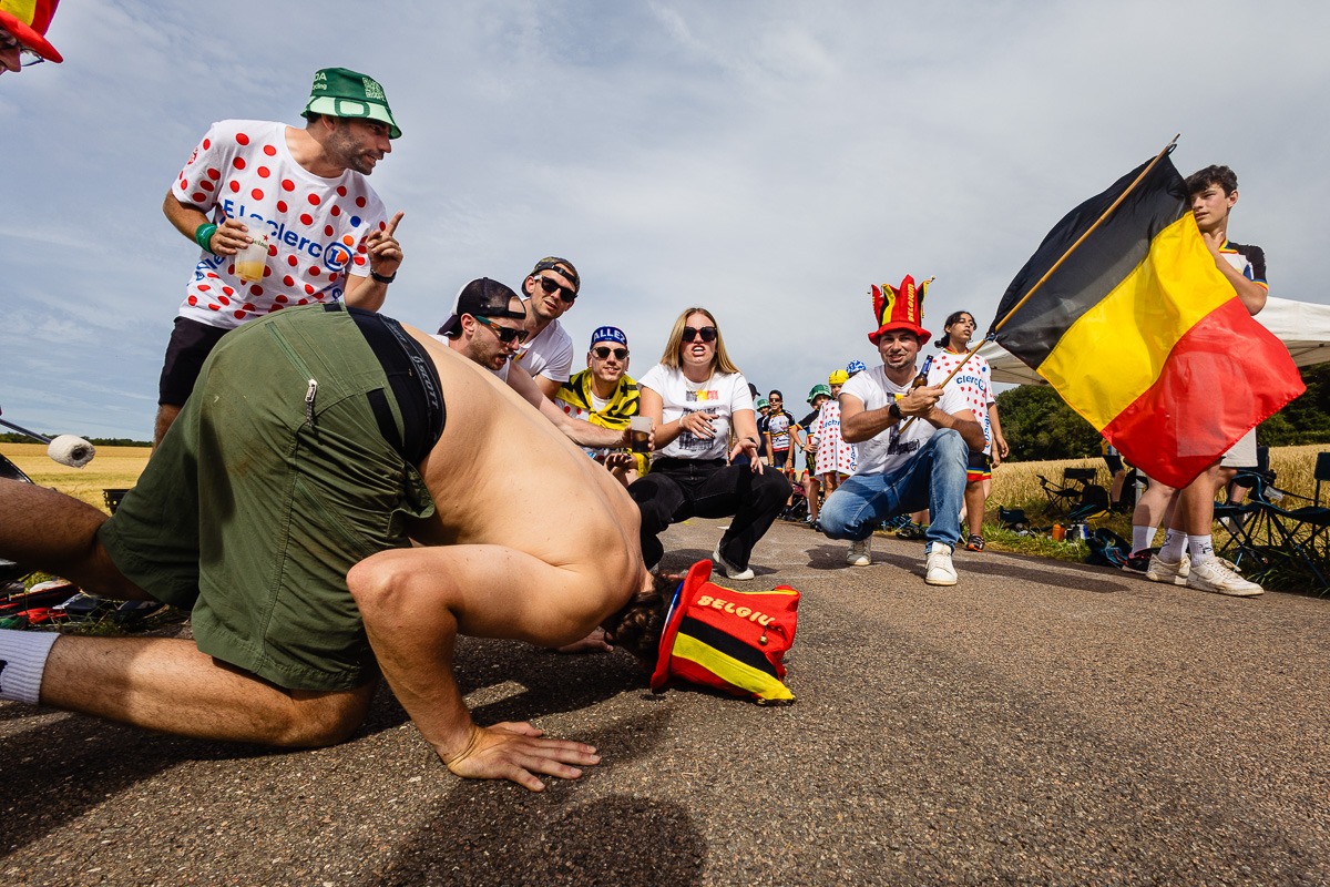 Gruppe festlich gekleideter Personen, die um einen Mann mit nacktem Oberkörper, der auf einer Straße Liegestütze macht, stehen und klatschen. Sie schwenken belgische Flaggen und scheinen zu feiern.