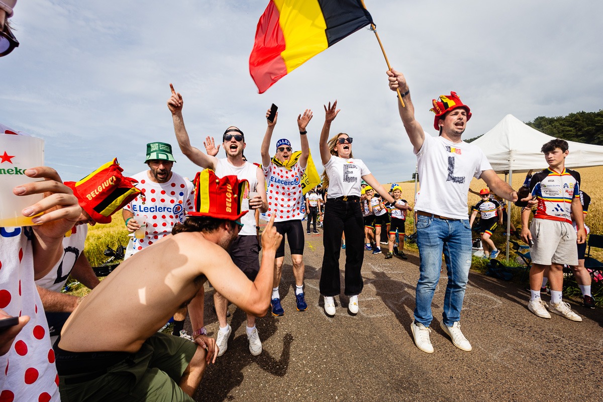 Eine Gruppe begeisterter Zuschauer klatscht und schwenkt bei einer Radsportveranstaltung eine belgische Flagge. Einige tragen gepunktete Trikots und lustige Hüte, und eine Person hält eine Tasse mit einem Getränkelogo.