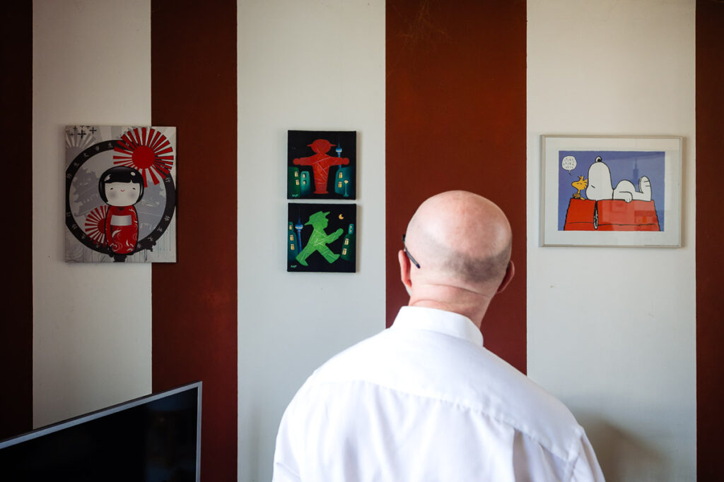 Un homme chauve en chemise blanche se tient face à un mur avec quatre œuvres d'art colorées de style dessin animé affichées entre des bandes verticales rouges et blanches.