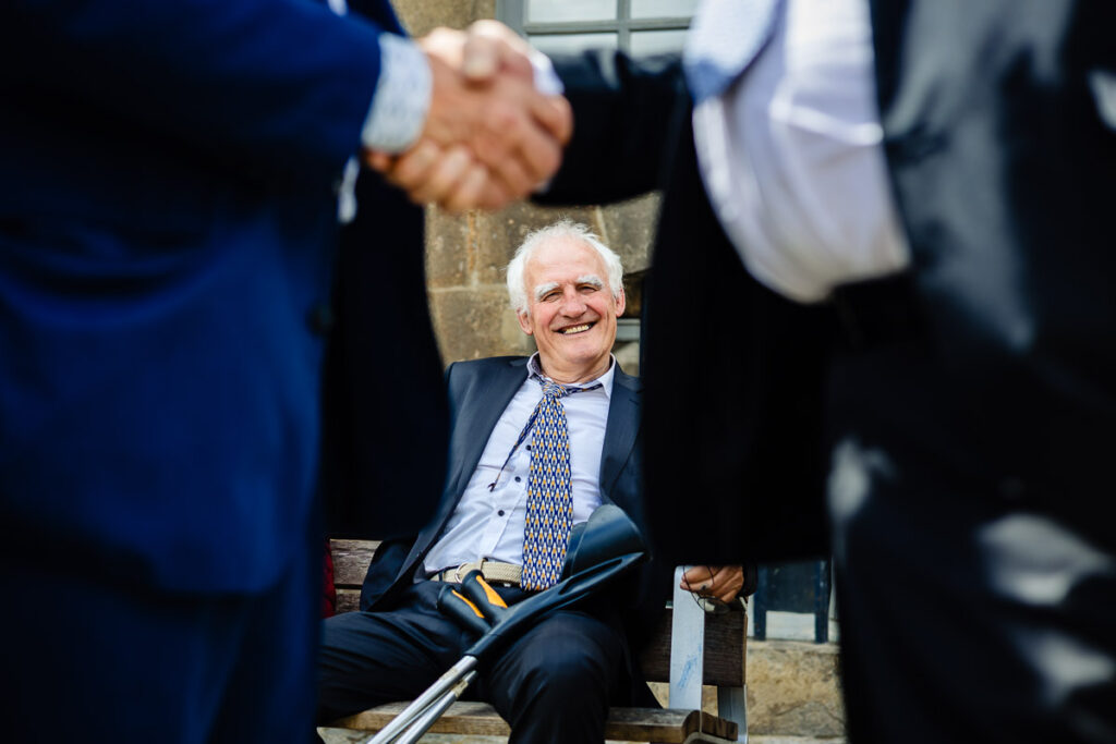 Un homme âgé en costume sourit assis sur un banc, avec deux personnes en costume se serrant la main au premier plan.