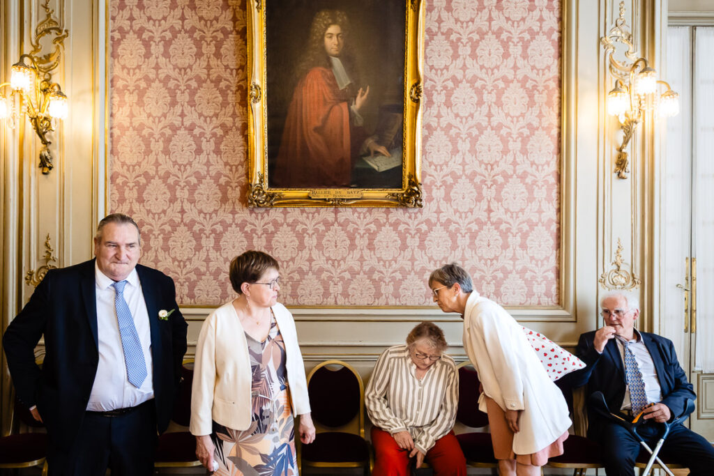 Cinq personnes âgées en tenue de soirée se réunissent dans une pièce décorée avec du papier peint à motifs et un grand portrait accroché au mur au-dessus d'eux.