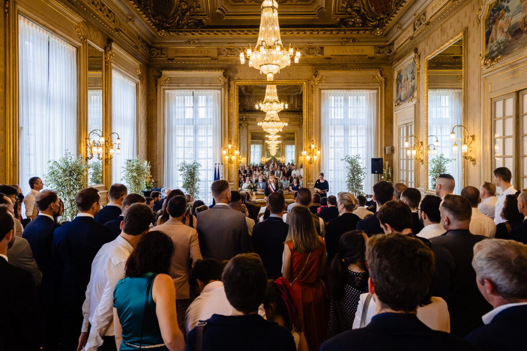 Un grand groupe de personnes se tient dans une pièce décorée, éclairée par des lustres et dotée de hautes fenêtres, écoutant un orateur à l'avant.