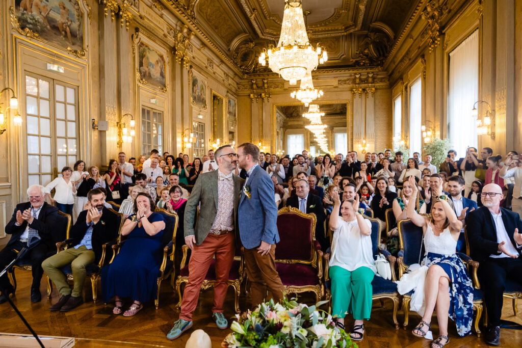 Deux hommes s'embrassent debout au centre d'une pièce richement décorée, entourés d'invités assis qui applaudissent et sourient lors d'une cérémonie de mariage.