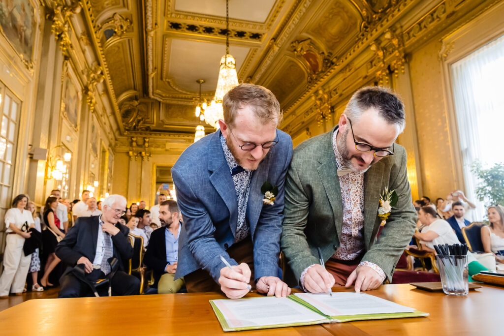 Deux hommes en costume signent des documents à une table dans une pièce décorée, tandis que les invités assis derrière eux regardent l'événement.