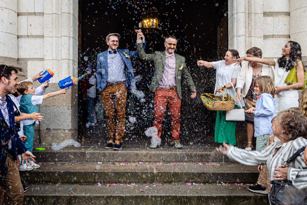 Deux hommes en costume se tiennent la main et descendent les marches de l'église tandis que les gens pulvérisent des bulles et jettent des confettis en signe de célébration.