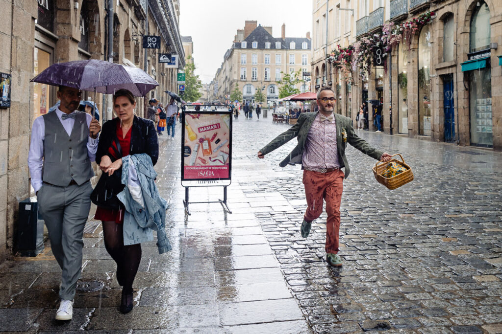 Deux personnes marchent sous un parapluie dans une rue pavée pluvieuse tandis qu'un homme portant un panier se précipite derrière eux, avec des magasins et des fleurs bordant la rue.