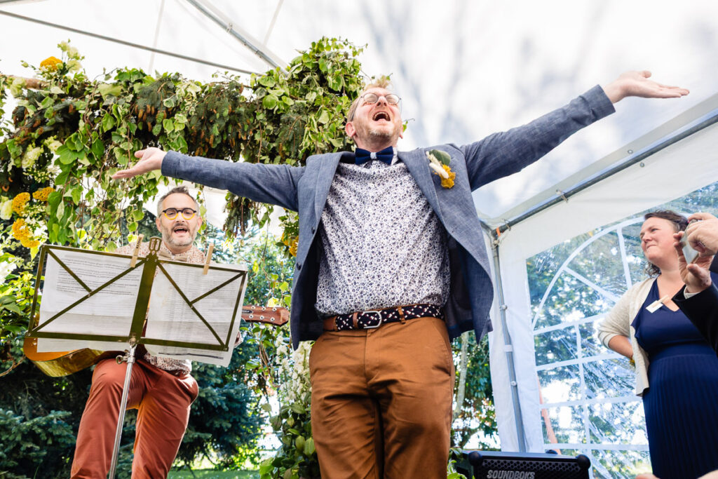 Un homme se tient debout, les bras tendus et la bouche ouverte, l'air joyeux, tandis qu'une autre personne joue de la guitare à côté de lui dans un décor d'événement en plein air décoré de verdure.