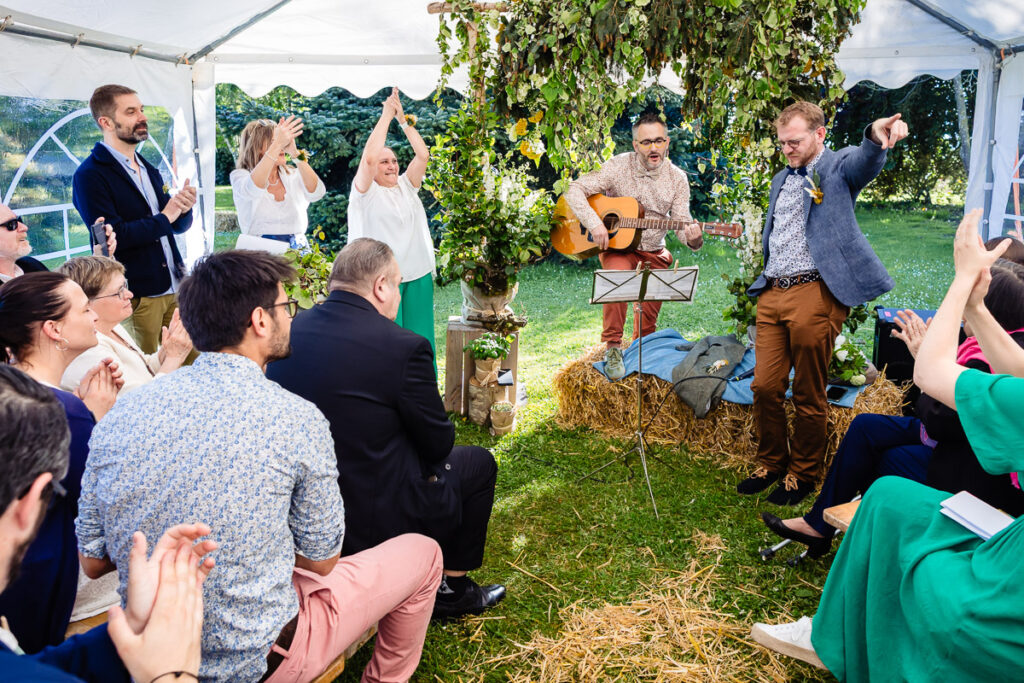Un groupe de personnes est assis et applaudit tandis que deux hommes jouent de la musique, l'un jouant de la guitare, sous une tente décorée de verdure et de balles de foin dans un décor extérieur.