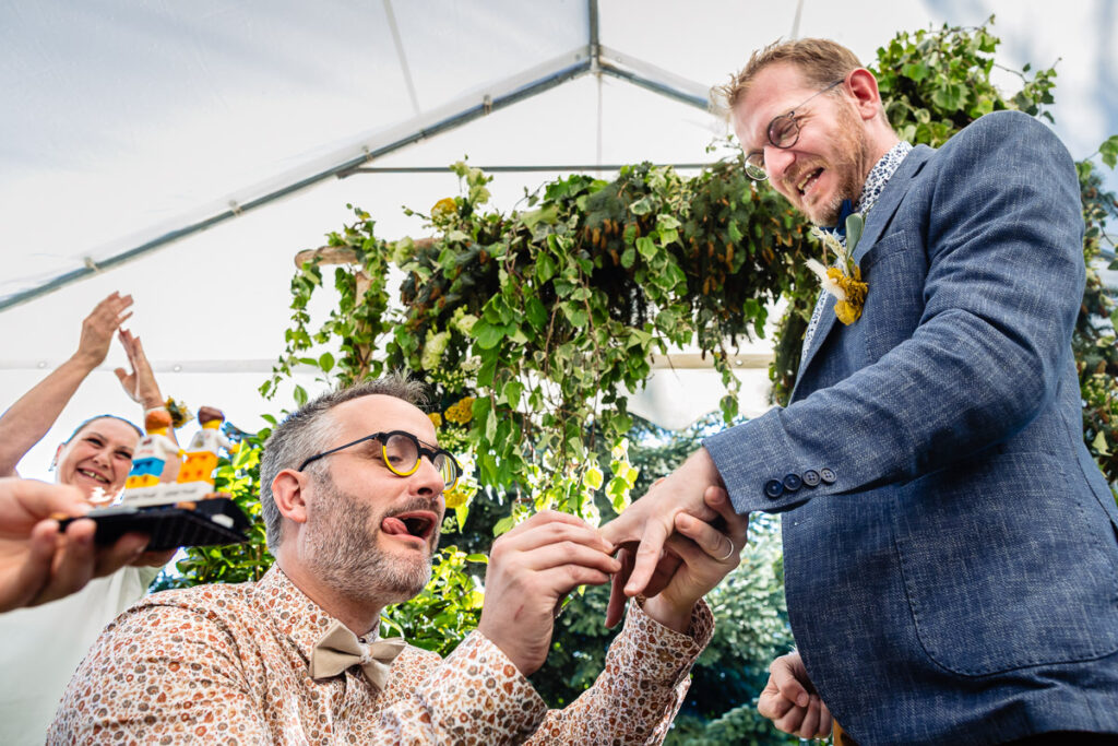 Deux hommes échangent des alliances lors d'une cérémonie de mariage en plein air sous une tente, tandis qu'une autre personne applaudit en arrière-plan au milieu de la verdure et des fleurs.