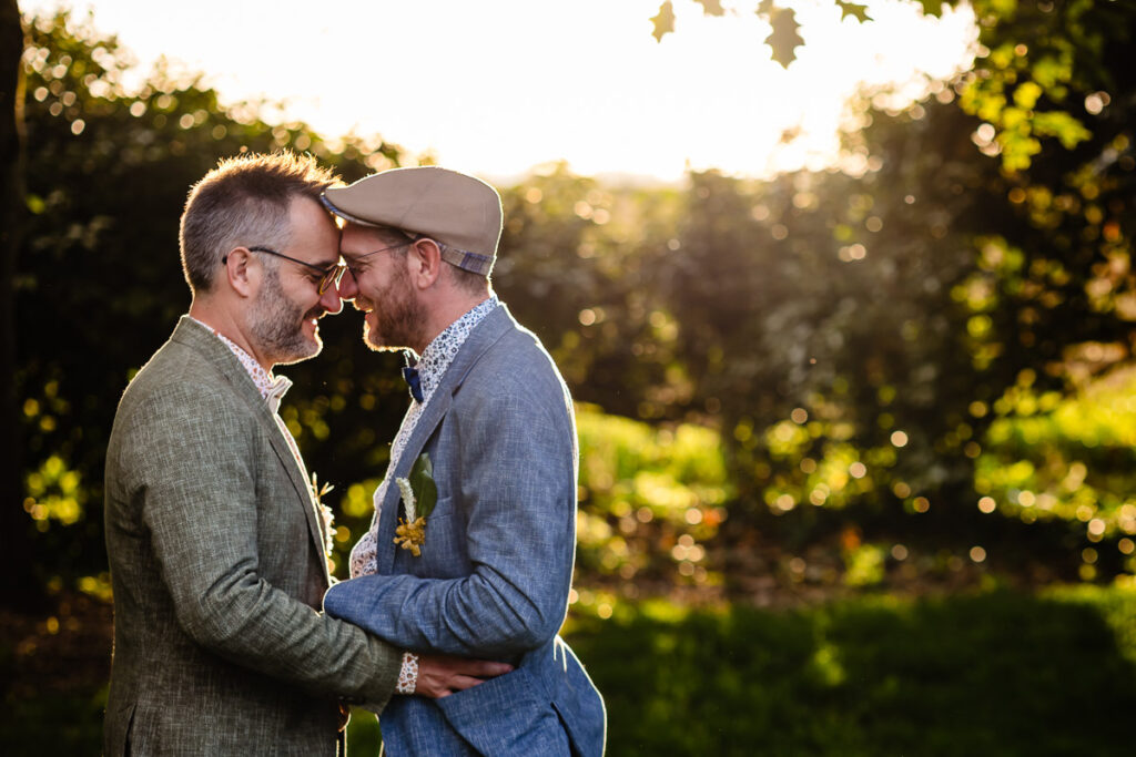 Deux hommes se tiennent dehors, face à face, souriant, leurs fronts se touchant, se tenant la main dans un cadre verdoyant et ensoleillé.