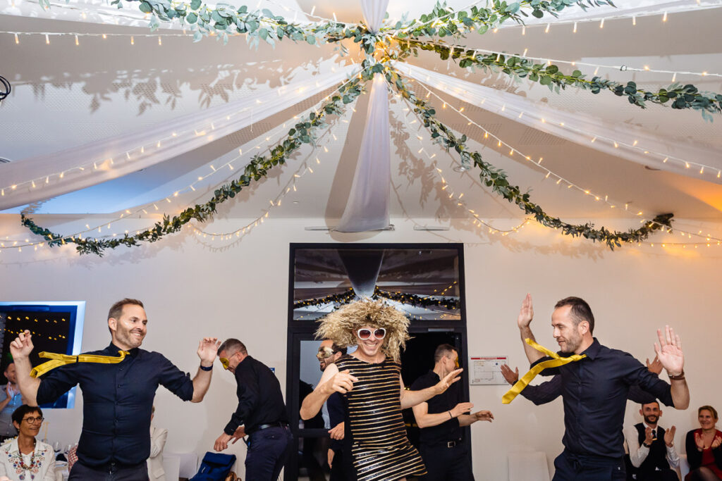 Une personne vêtue d'une robe rayée, d'une grande perruque et de lunettes de soleil danse énergiquement avec trois hommes en chemises sombres et cravates jaunes sous des lumières décoratives lors d'un événement festif en salle.