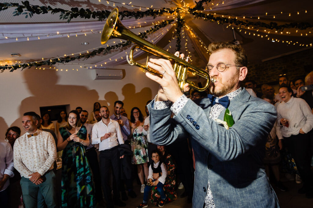 Un homme en costume joue de la trompette lors d'un événement en salle, entouré d'une foule de personnes sous des guirlandes lumineuses et des décorations au plafond.