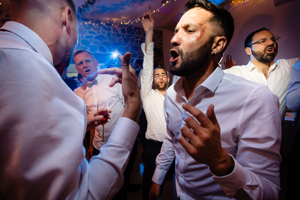 Un groupe d'hommes en chemises blanches chantant et gesticulant avec enthousiasme lors d'une fête intérieure animée avec des éclairages festifs.