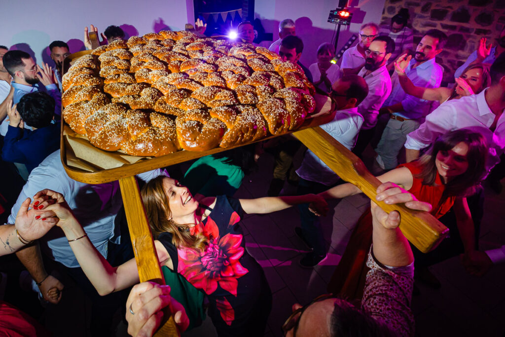 Les gens soulèvent un gros pain tressé garni de graines sur un plateau en bois lors d'un rassemblement intérieur animé.