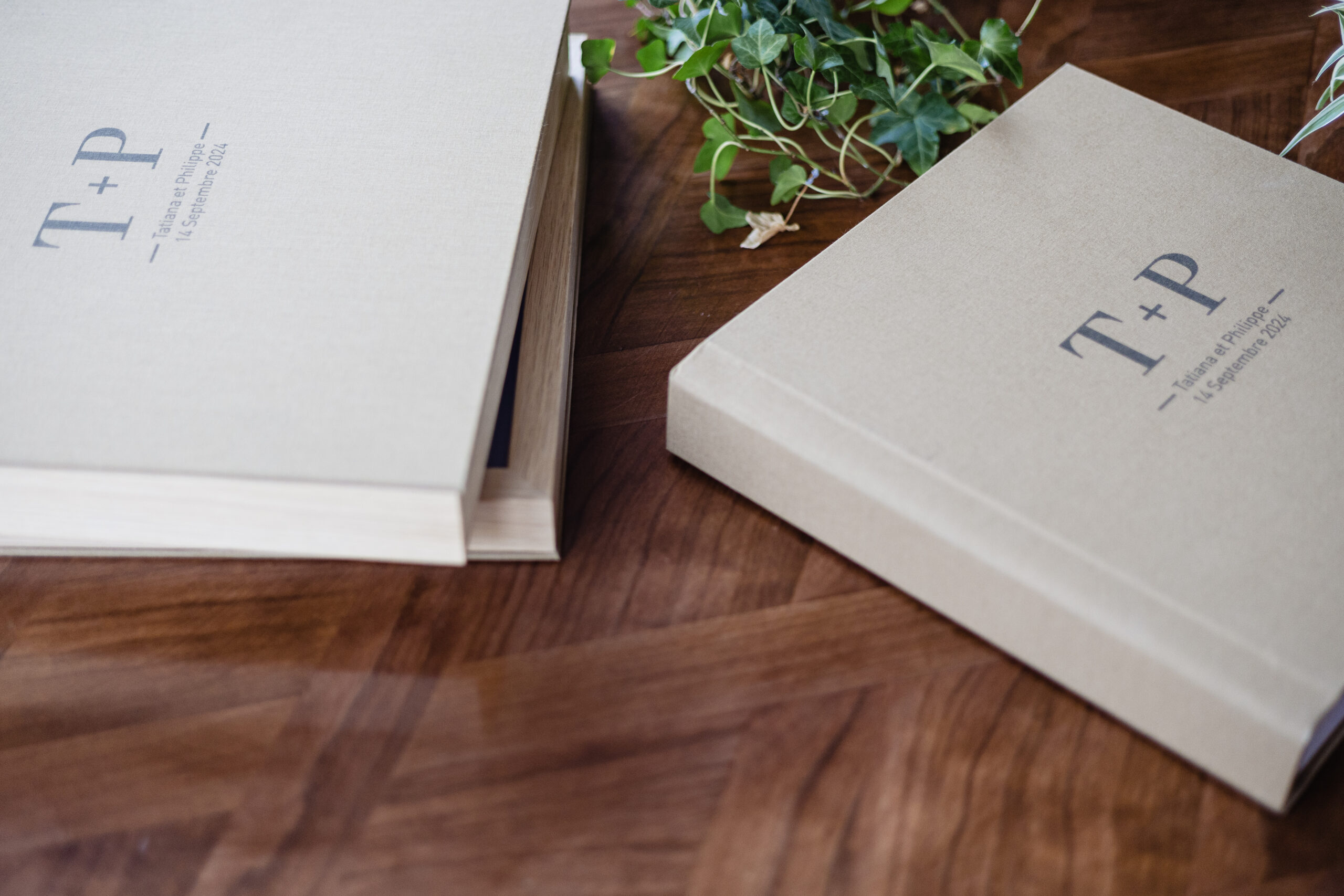 Deux albums beiges à couverture rigide portant la mention "T + P 14 février 2023" reposent sur une surface en bois à proximité de quelques plantes à feuilles vertes, parfaites pour conserver vos tirages préférés.