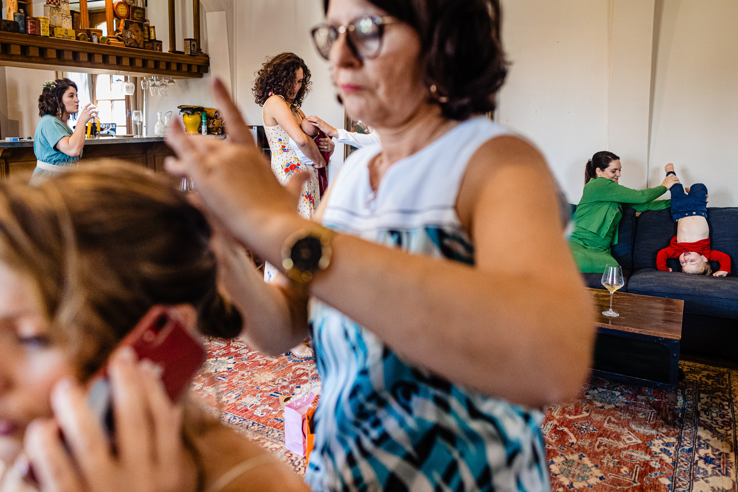 Une femme coiffe une autre femme tout en parlant au téléphone ; à l'arrière-plan, deux femmes conversent près d'un comptoir et un enfant est allongé à l'envers sur un canapé.