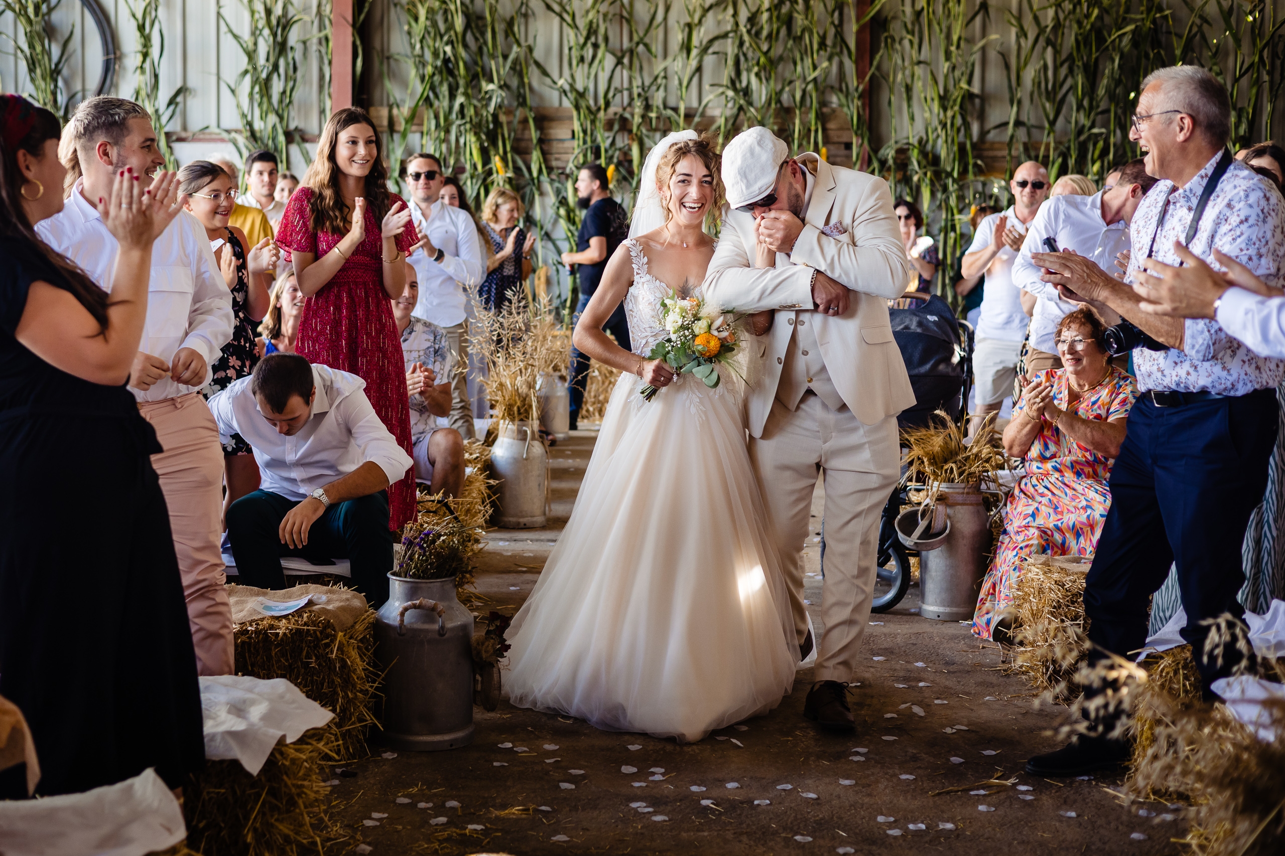 Une mariée en robe blanche descend l'allée avec un homme plus âgé en costume blanc, entourée d'invités assis et debout qui applaudissent dans un lieu rustique et décoré.