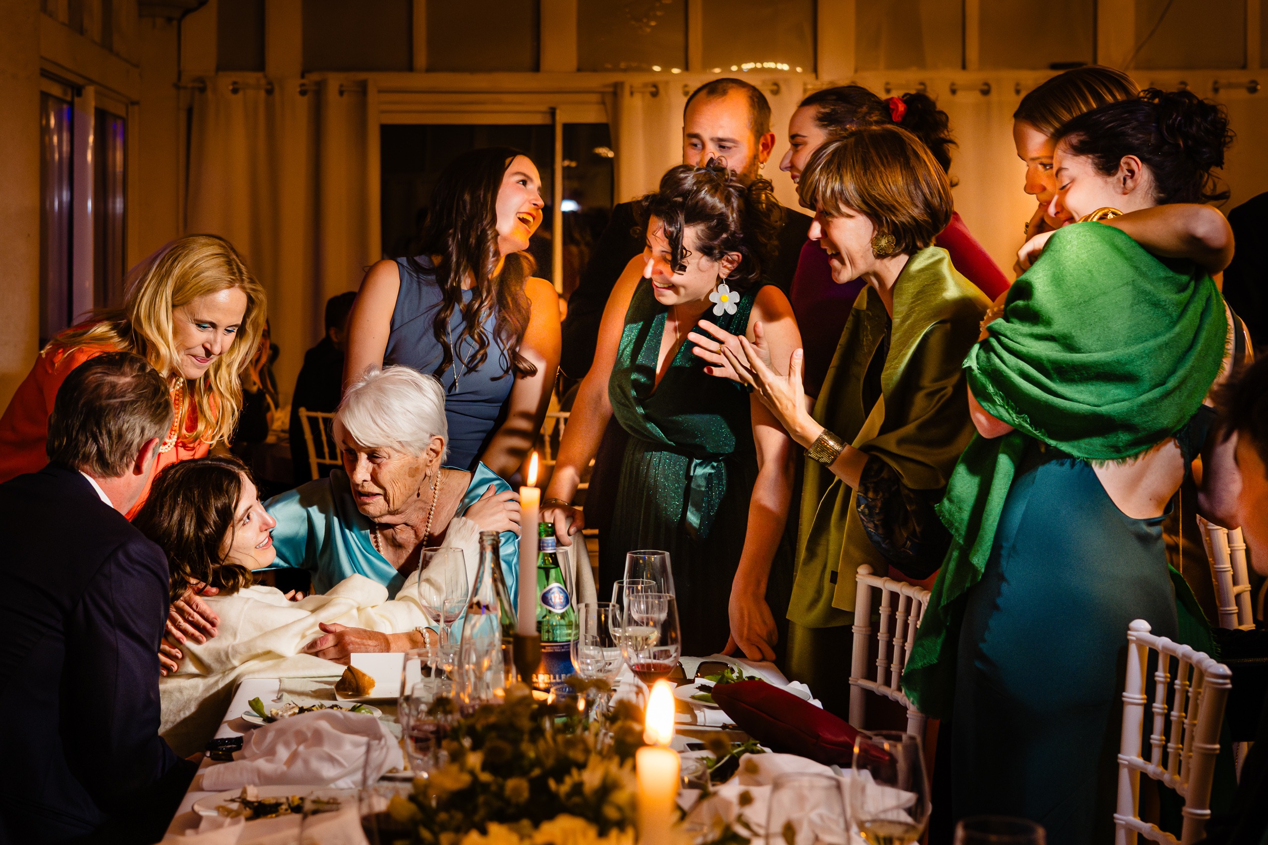 Un groupe de personnes, principalement des femmes, se rassemblent autour d'une femme âgée assise à une table de dîner garnie de verres, d'assiettes et de bougies, dans un intérieur chaleureusement éclairé.