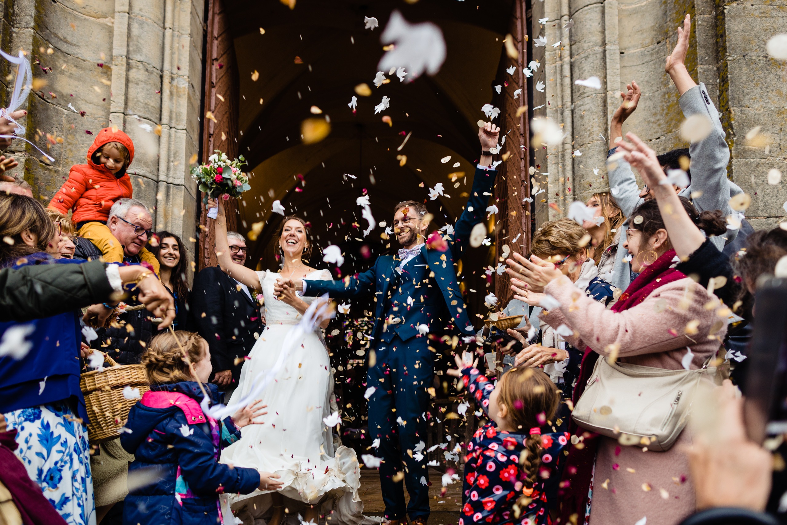 Des mariés marchent à l'extérieur d'une église tandis que des invités les acclament et leur lancent des pétales de fleurs pour célébrer leur mariage.