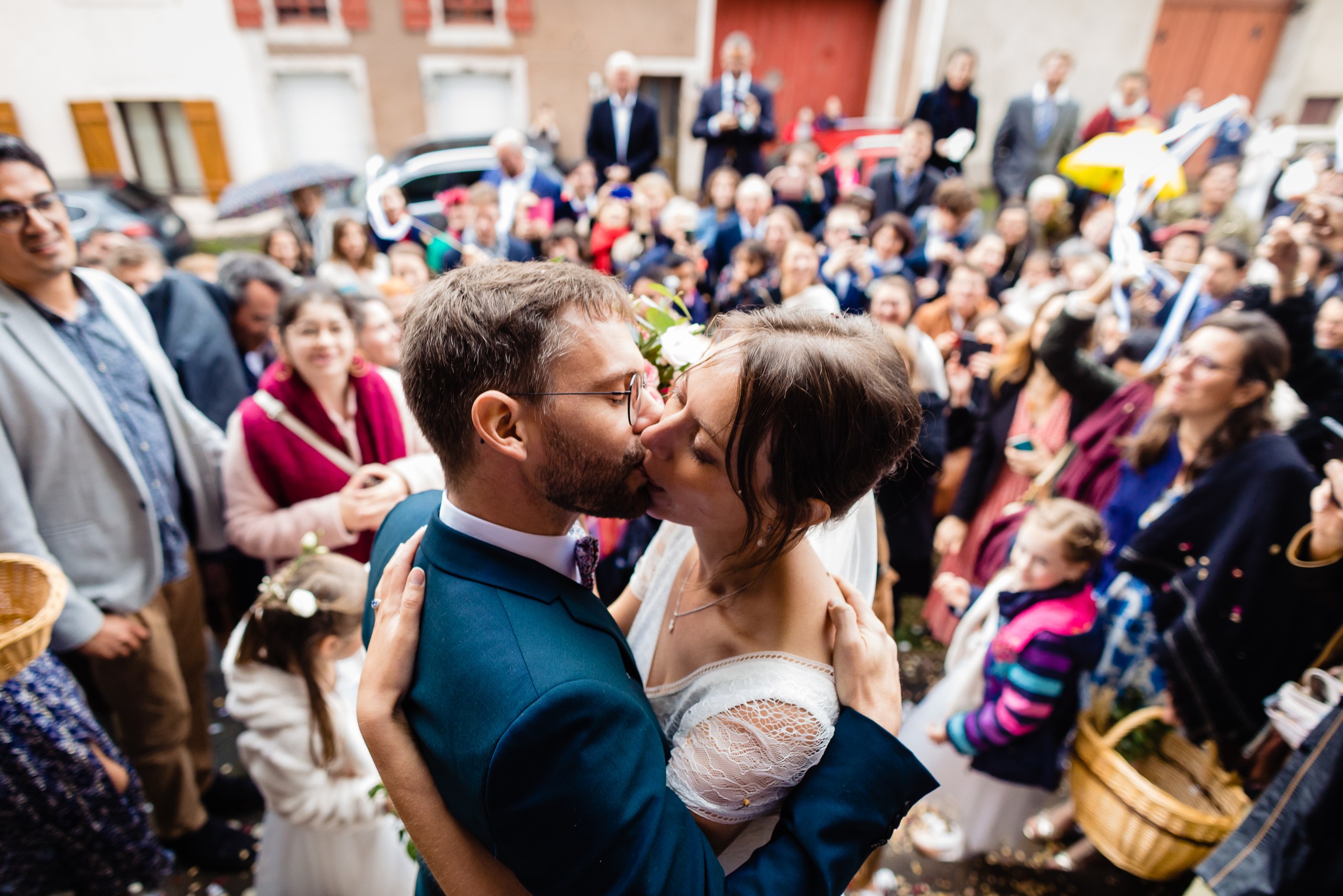 Des mariés s'embrassent en plein air, entourés d'une foule d'invités, dont certains sourient et regardent, lors de ce qui semble être une célébration de mariage.