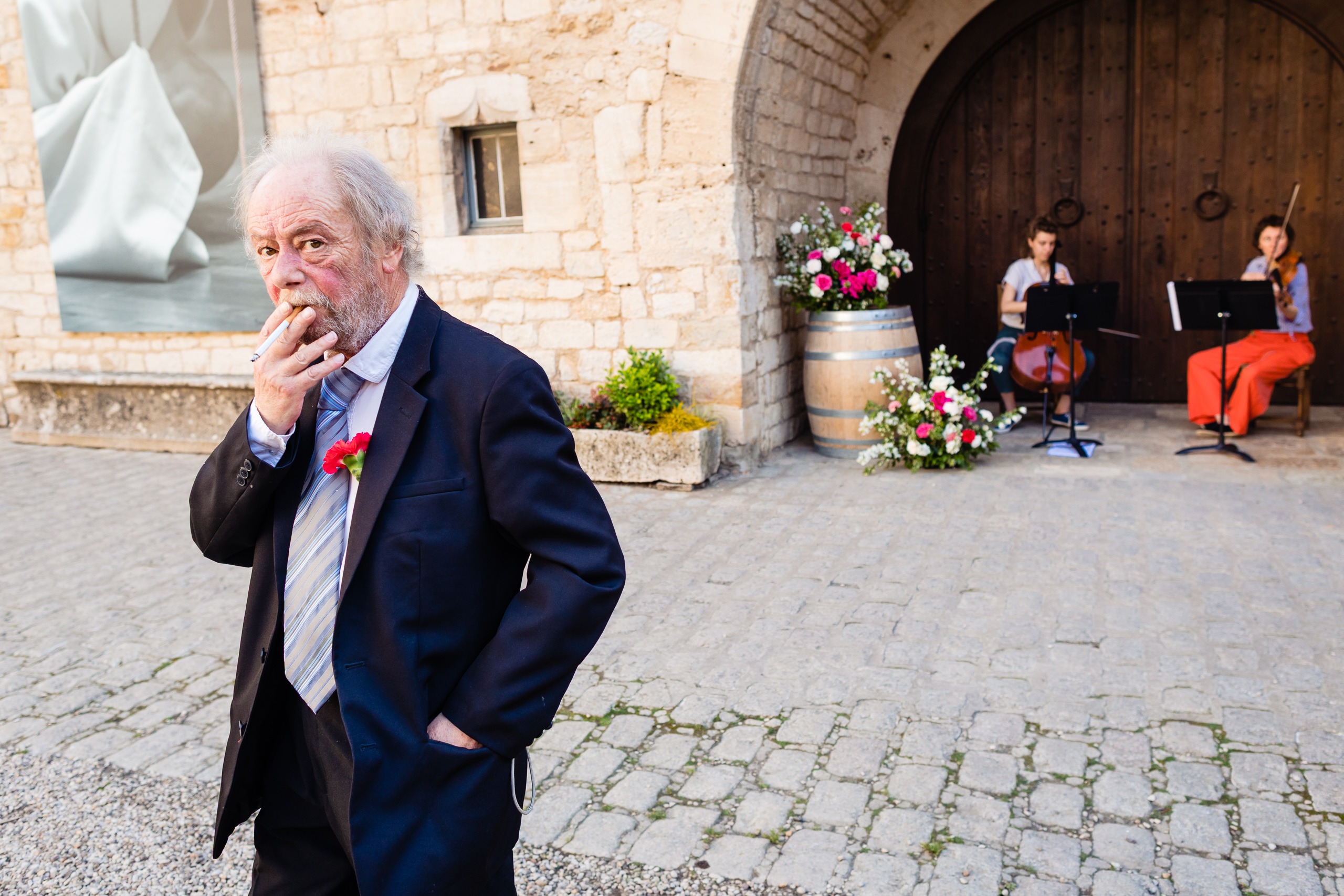 Un homme âgé en costume fume une cigarette à l'extérieur d'un bâtiment en pierre, tandis que deux musiciens jouent des instruments à cordes près d'une grande porte en bois ornée de compositions florales.