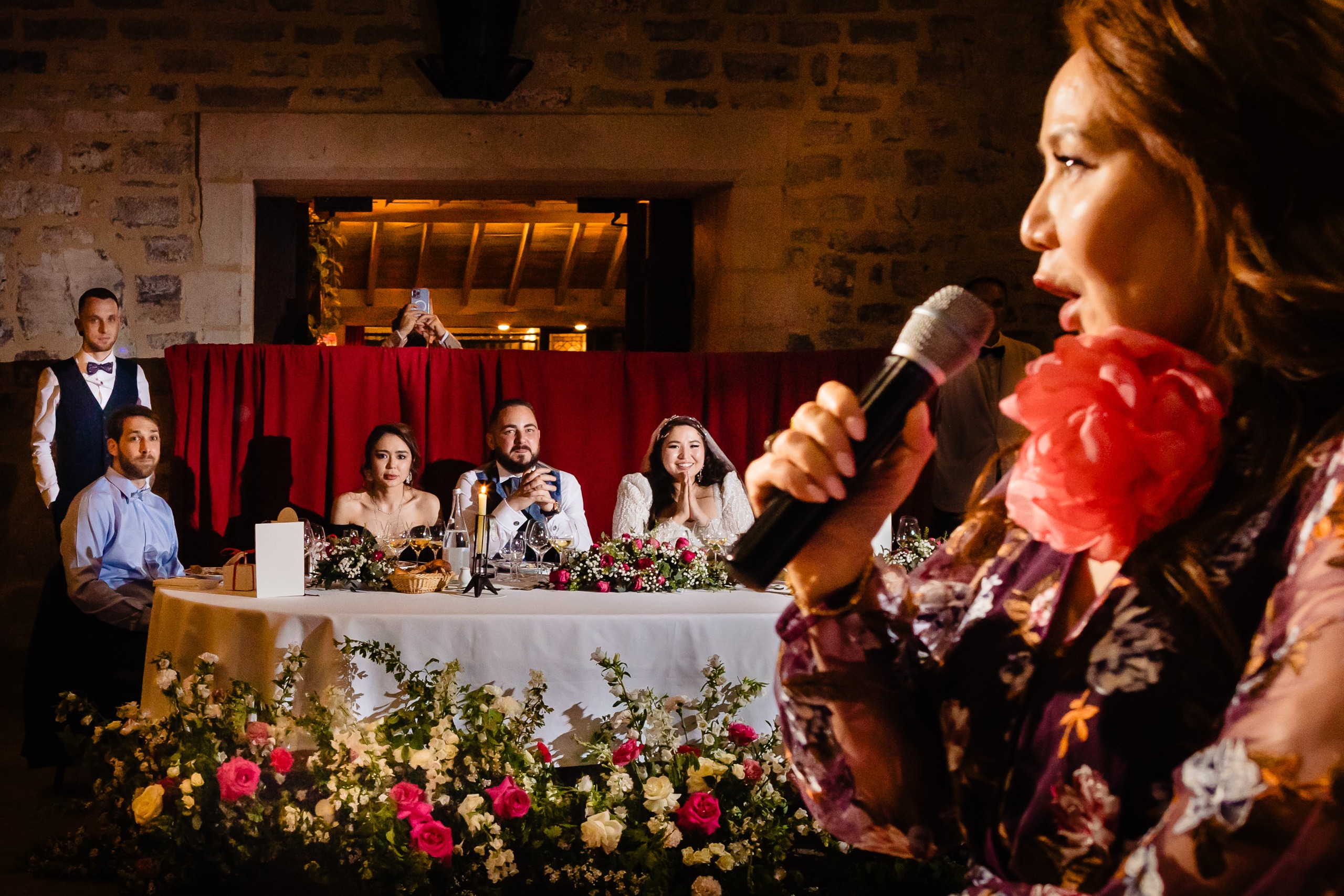 Une femme parle dans un microphone lors d'une réception de mariage, tandis que la mariée, le marié et les invités sont assis à une table décorée à l'arrière-plan.