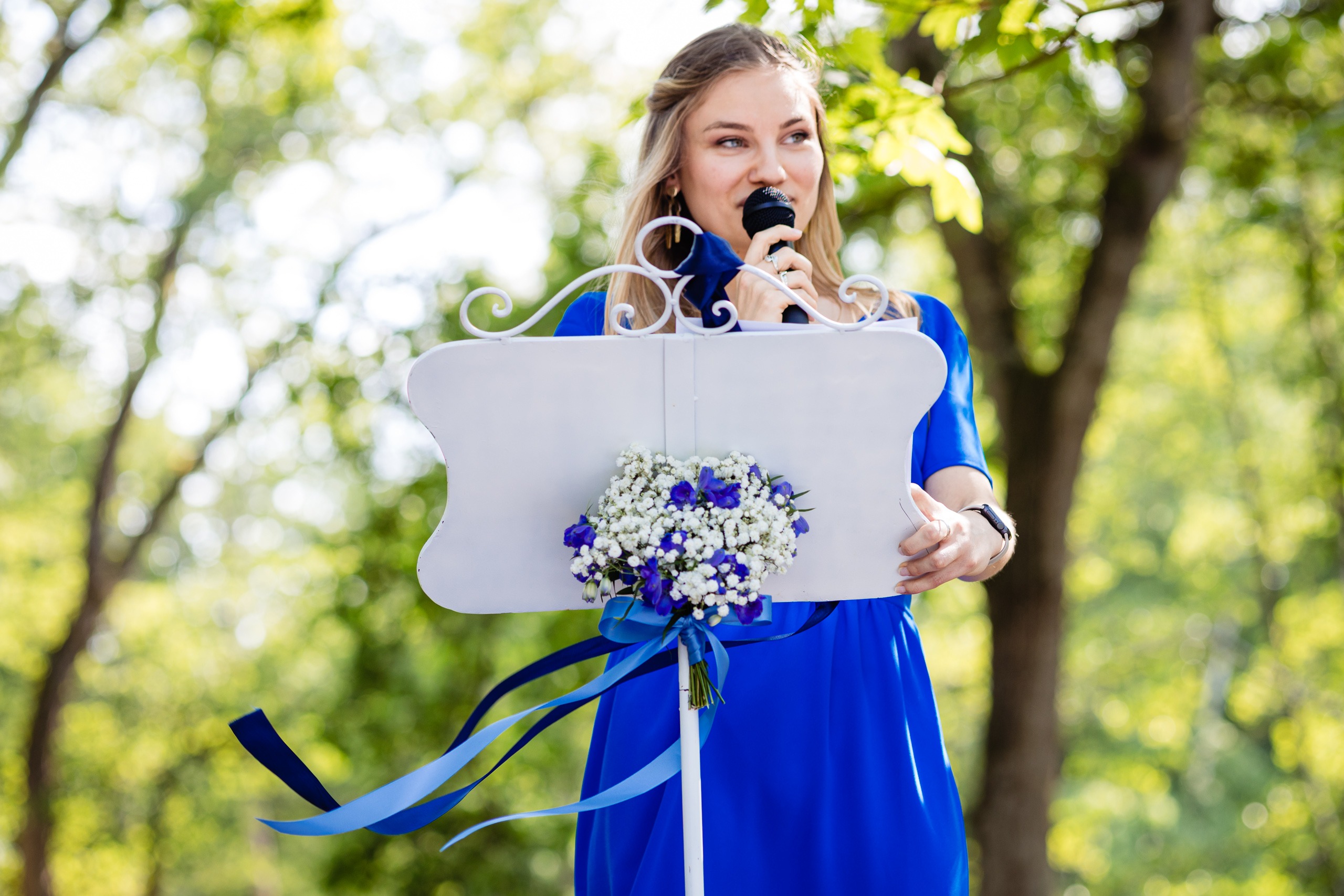 Une femme vêtue d'une robe bleue parle dans un microphone sur un podium extérieur décoré de fleurs et de rubans blancs et bleus. Des arbres sont visibles à l'arrière-plan.