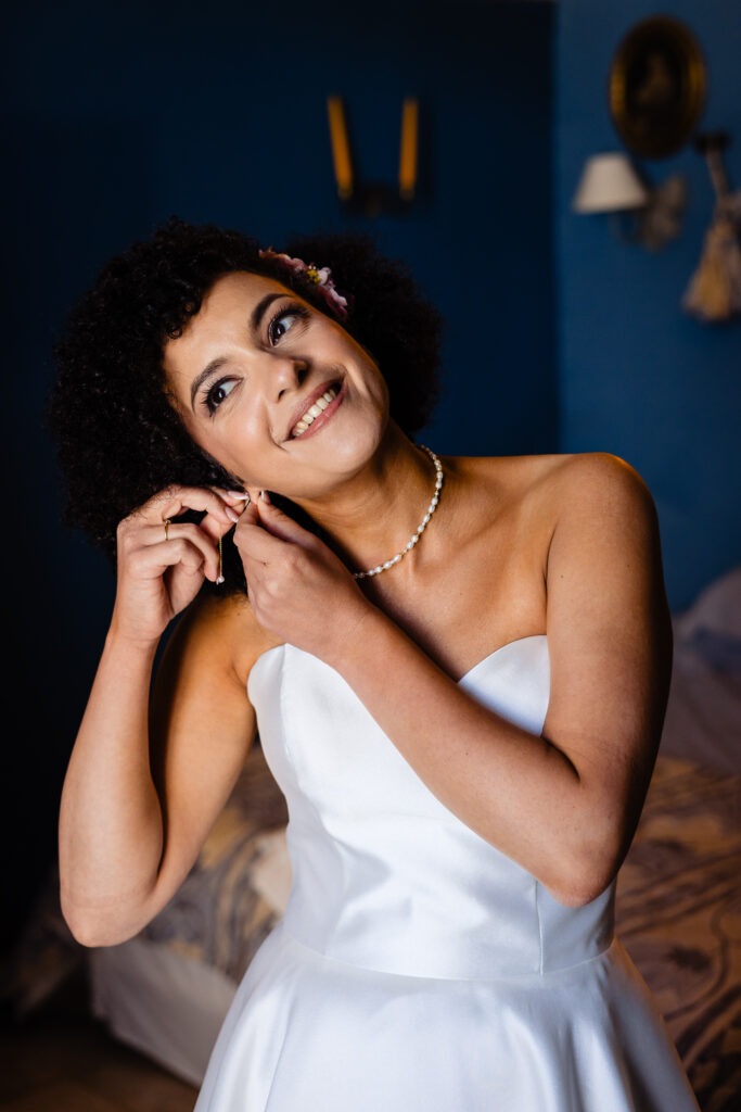 Une femme en robe bustier blanche sourit en mettant une boucle d'oreille, debout dans une pièce aux murs bleus le jour de son mariage, habilement capturé par un photographe de Dijon.