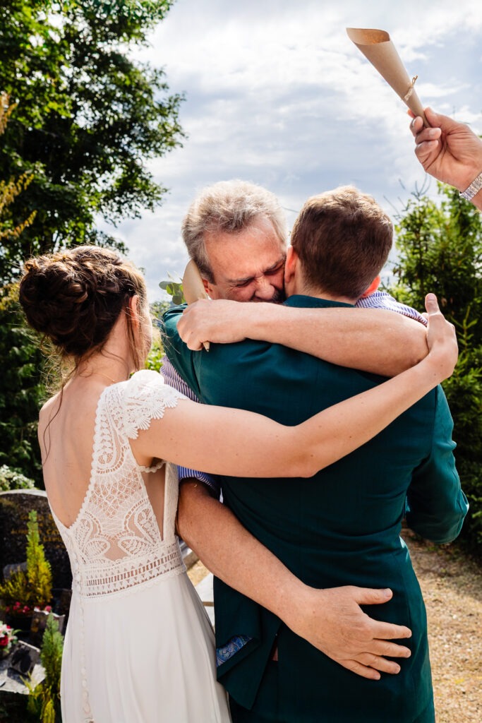 Les mariés se serrent dans les bras d'un homme plus âgé en plein air, tandis que leur mariage est magnifiquement capturé par un photographe à Dijon, avec une autre personne tenant un papier enroulé à l'arrière-plan.