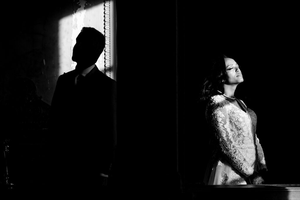 Une femme en robe blanche se tient dans la lumière du soleil, les yeux fermés, tandis qu'un homme en costume se tient dans l'ombre à proximité - une photo prise par un photographe mariage Dijon.