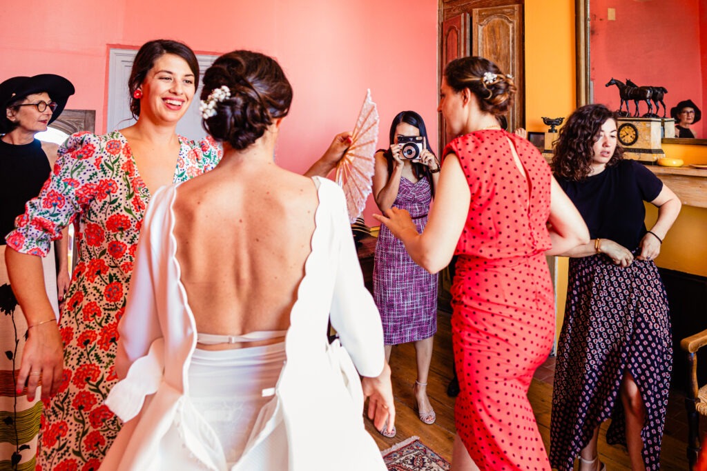 Une mariée en robe blanche se tient parmi des femmes en robes colorées, l'une d'entre elles tenant un appareil photo, dans une pièce lumineuse aux murs orange et jaunes lors d'un mariage à Dijon, capturé par le photographe.