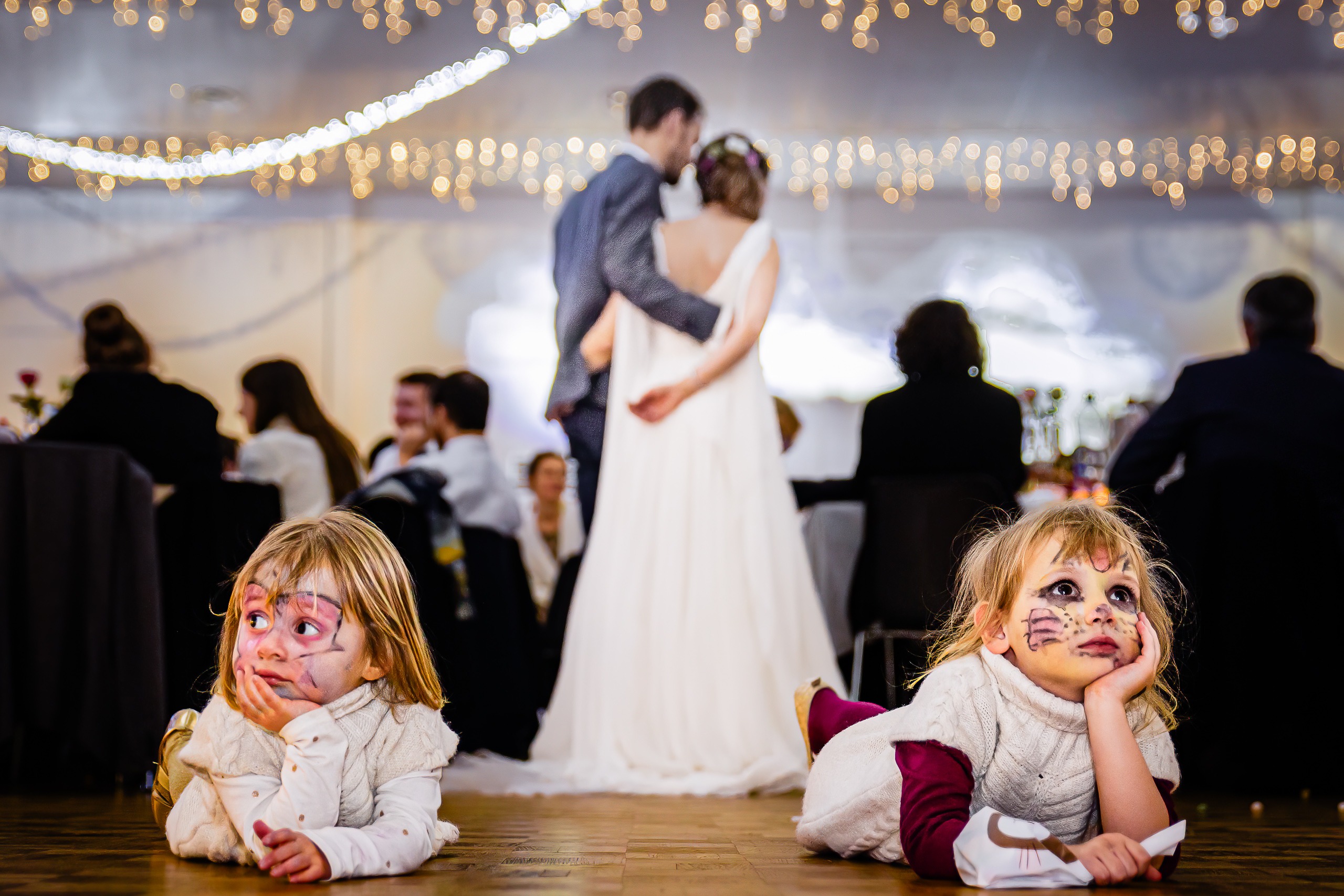 Deux jeunes enfants maquillés sont allongés sur le sol lors d'une réception de mariage, tandis qu'une mariée et un marié se tiennent ensemble dans un arrière-plan flou.
