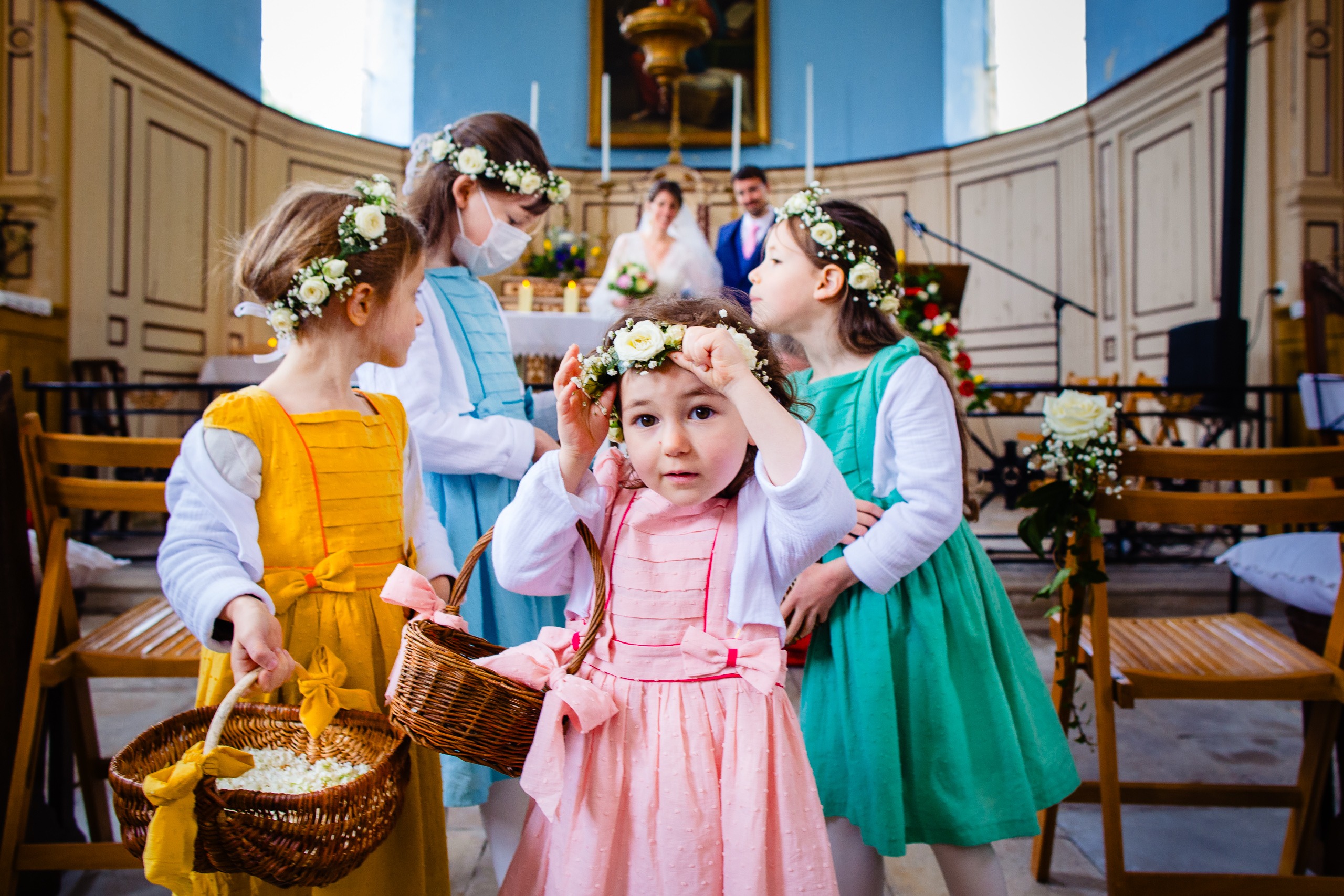 Cinq jeunes bouquetières en robes colorées se tiennent au premier plan d'une église, ajustant des couronnes de fleurs, avec des mariés flous à l'arrière-plan.