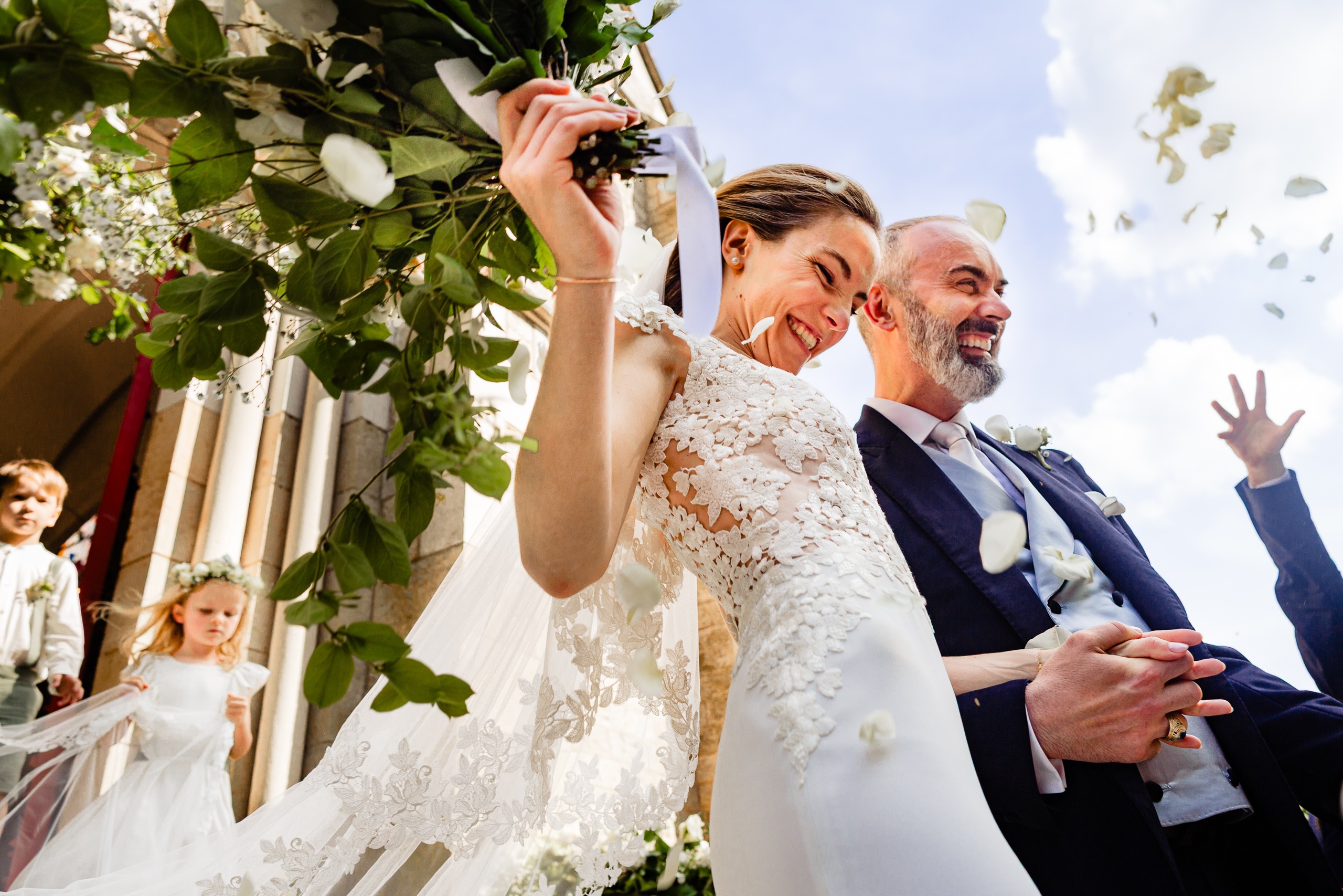Une mariée et un marié sourient et marchent à l'extérieur d'un bâtiment, se tenant par la main, entourés de pétales de fleurs et d'invités, dans une journée lumineuse et un ciel bleu.