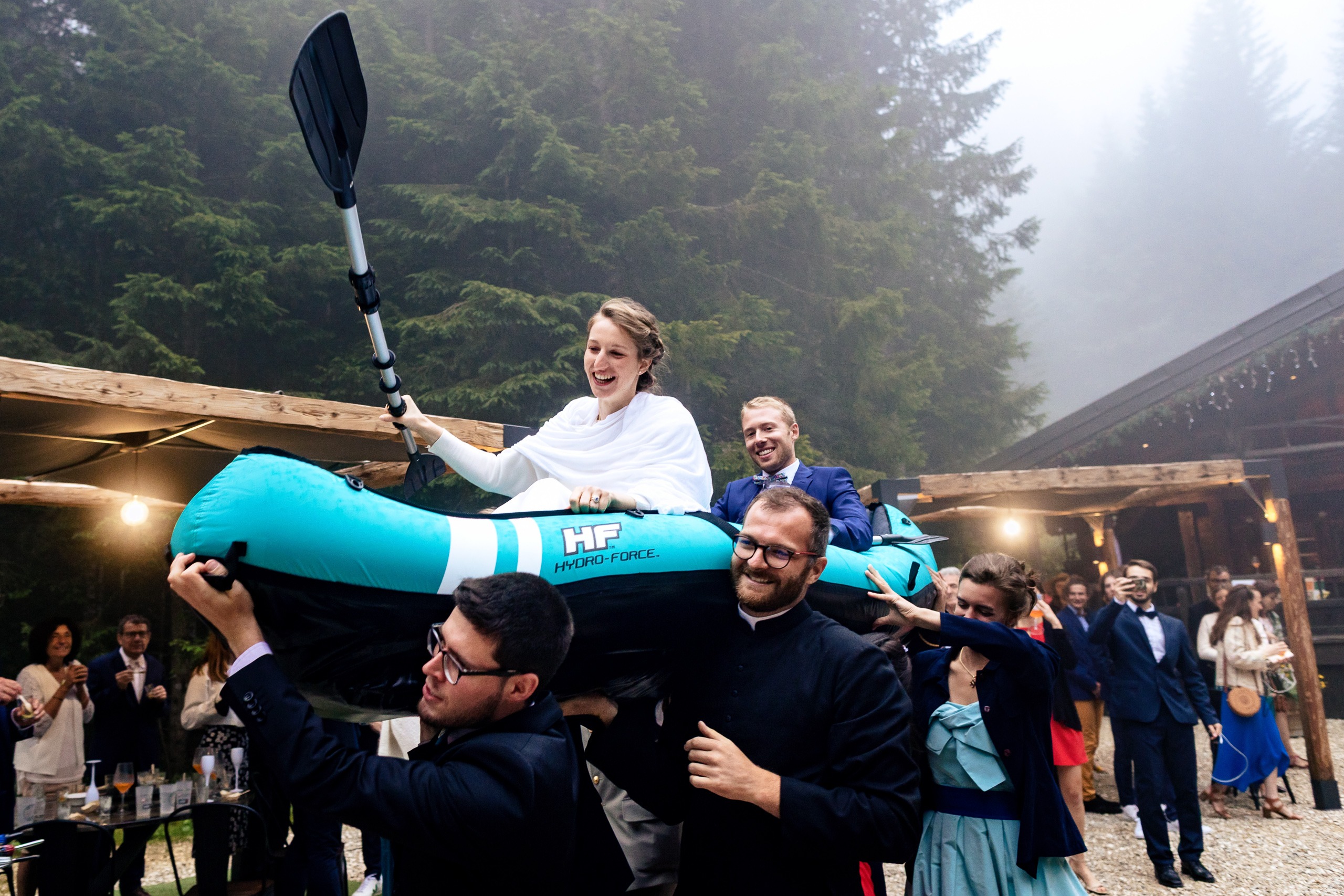 Les mariés sont assis dans un kayak gonflable tenu par des invités lors d'un mariage en plein air, avec des arbres et une structure en bois en arrière-plan.