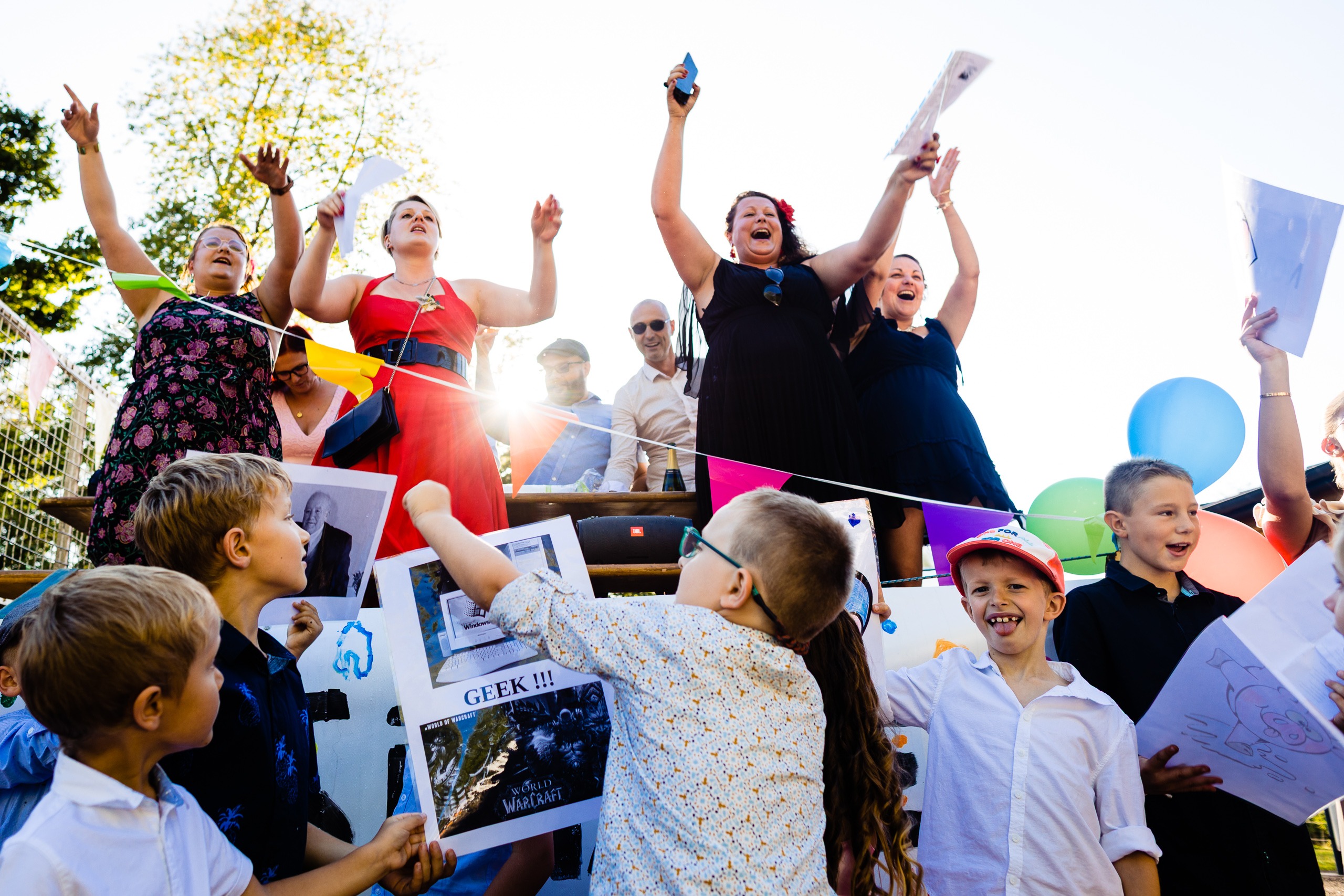 Un groupe d'enfants et d'adultes fêtent l'événement en plein air avec des pancartes, des ballons et des guirlandes ; certaines personnes applaudissent en levant les bras sous un soleil éclatant.