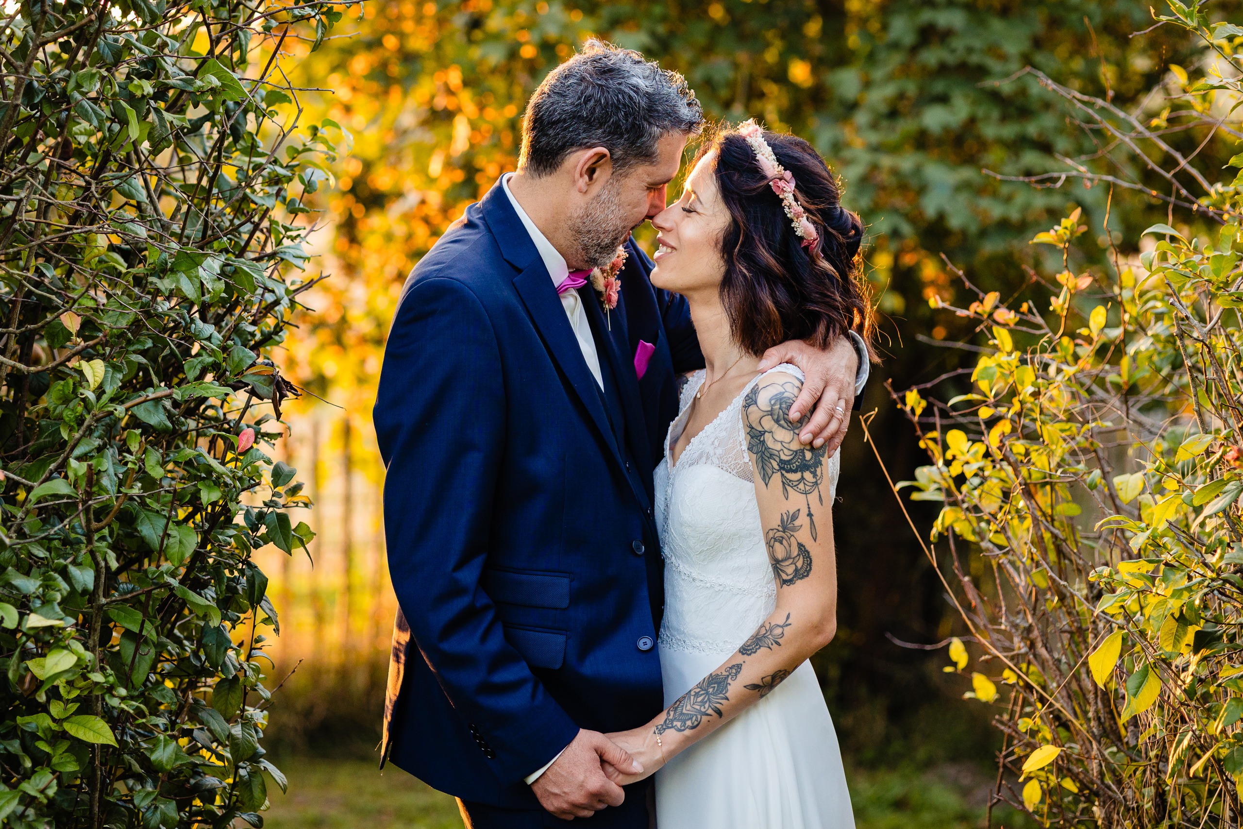 Un couple se tient debout à l'extérieur, se tenant par la main et se touchant le front. L'homme porte un costume bleu marine ; la femme porte une robe blanche et un bandeau fleuri. Ils sont entourés de feuillage vert.