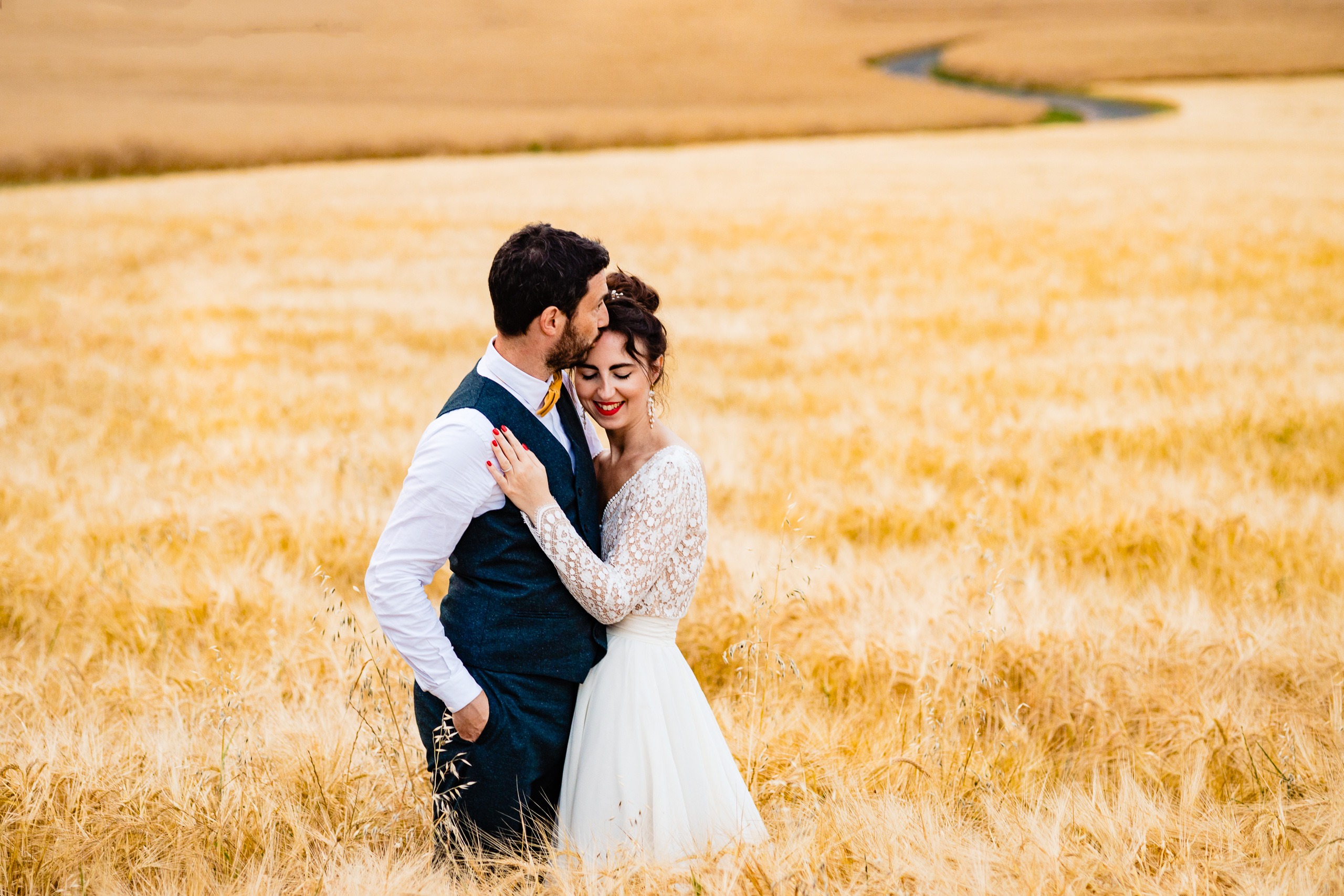 Un couple en tenue de mariage se tient enlacé dans un champ de blé doré, le marié embrassant le front de la mariée.