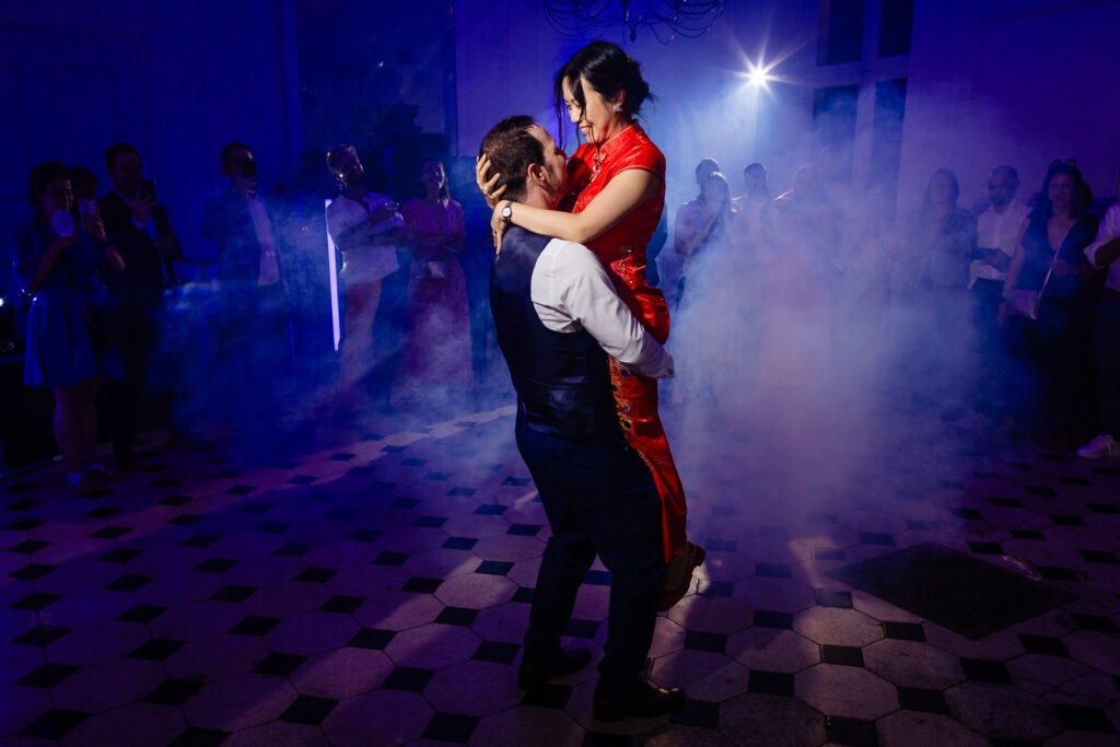 Un homme soulève une femme vêtue d'une robe rouge alors qu'ils dansent sur une piste à carreaux, entourés de gens et d'un éclairage de scène bleu avec des effets de fumée, photographiés par un photographe de mariage à Dijon.