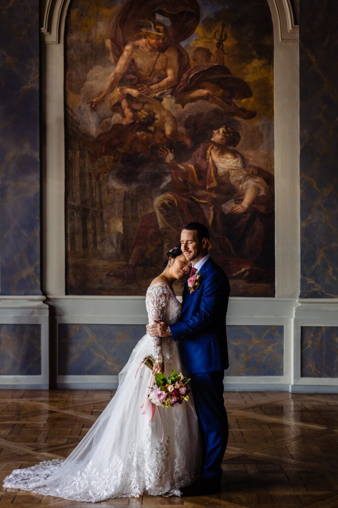 Les mariés sont debout, enlacés à l'intérieur, la mariée tenant un bouquet, avec un grand tableau classique à l'arrière-plan. Cette photo a été prise par un photographe mariage de Dijon.